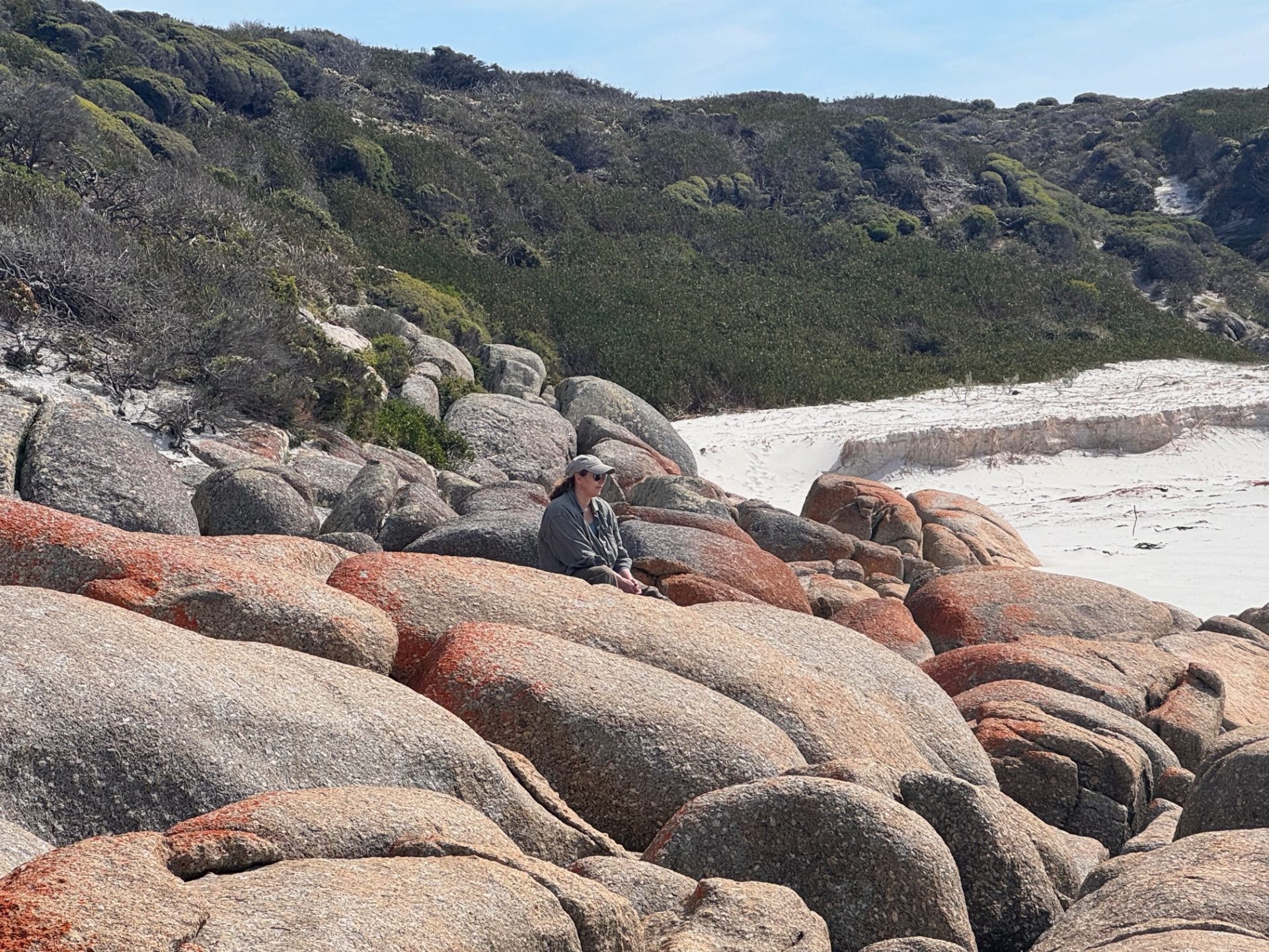 Photo of a beach with large rocks in the foreground. A woman sits amongst the rocks, looking out to sea.