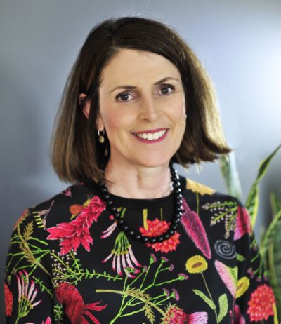 Professional headshot of a smiling white woman with straight dark hair, cut to a shoulder length bob. She is wearing a black top that has a bright floral pattern.